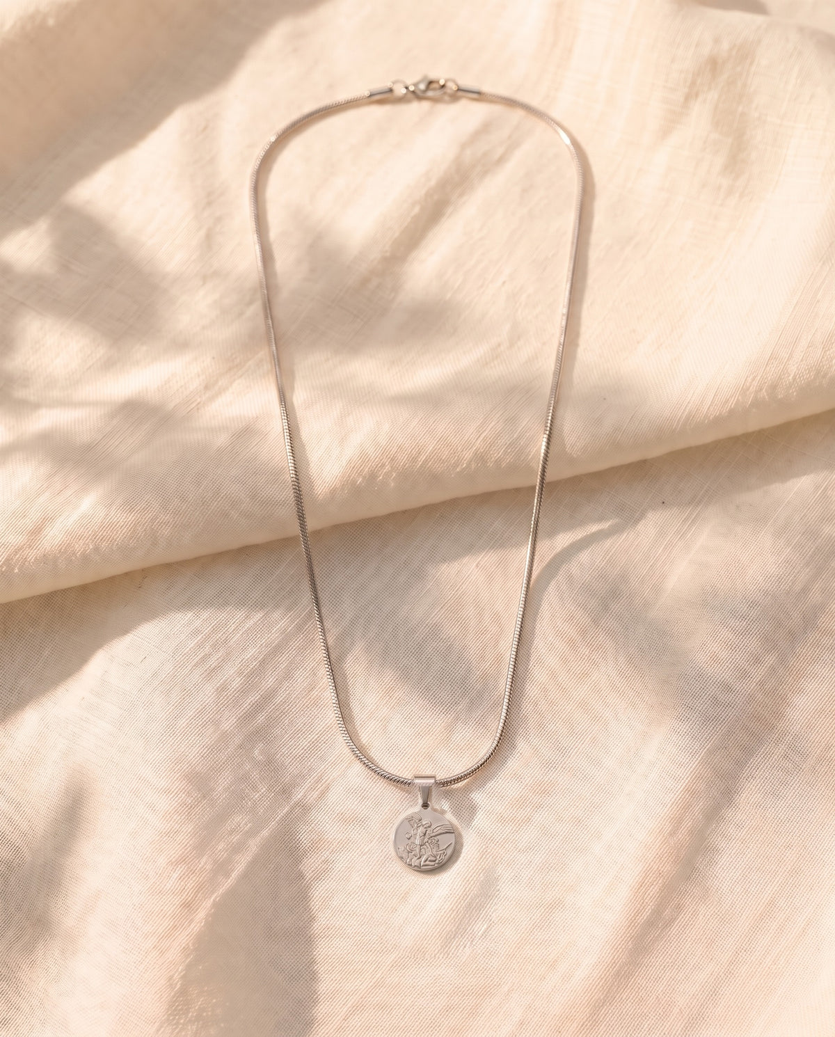 Marco Maveri Archangel Medallion Snake Chain Necklace laid on cream linen, Silver