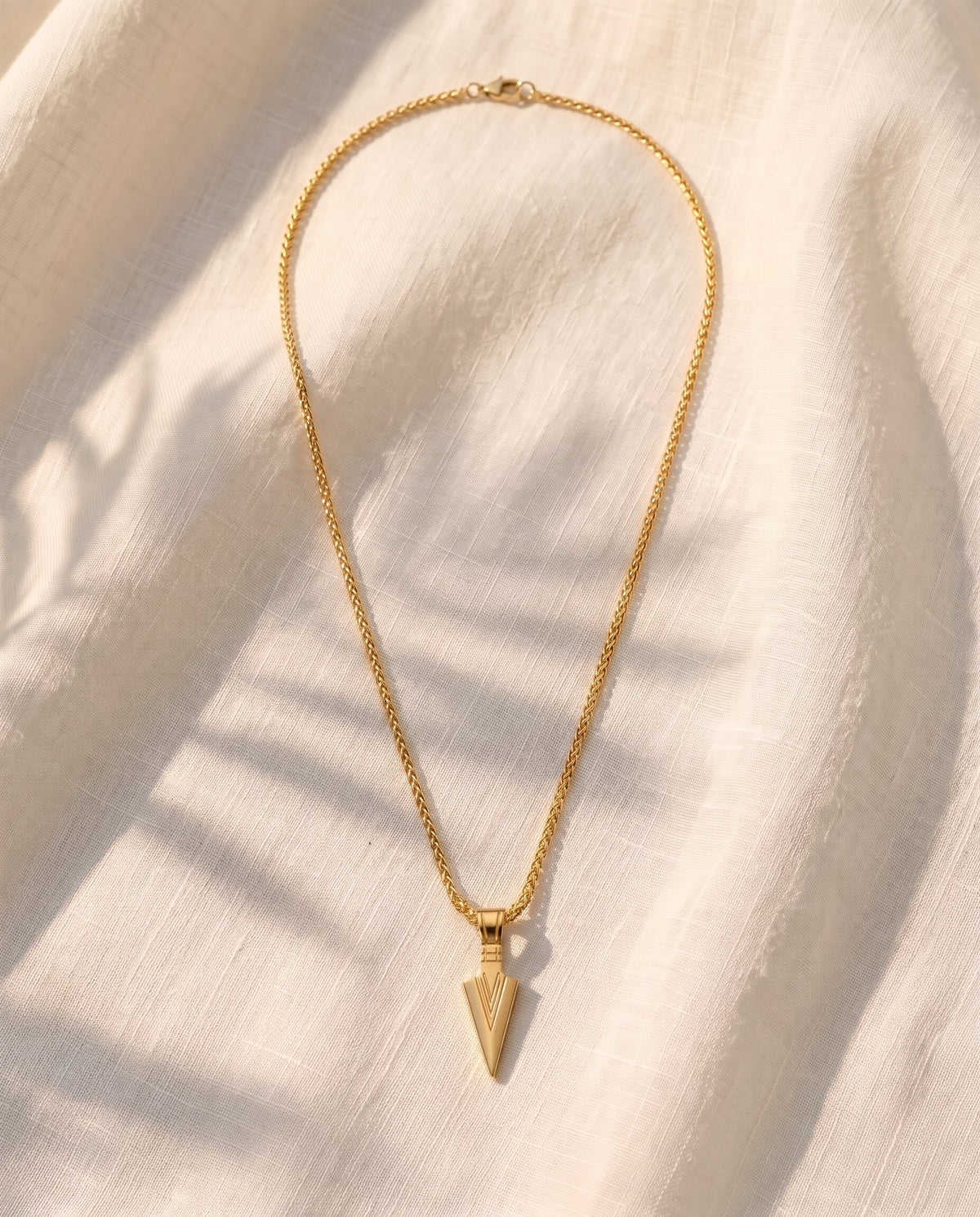 Marco Maveri Arrowhead Pendant Wheat Chain Necklace laid on cream linen, Gold