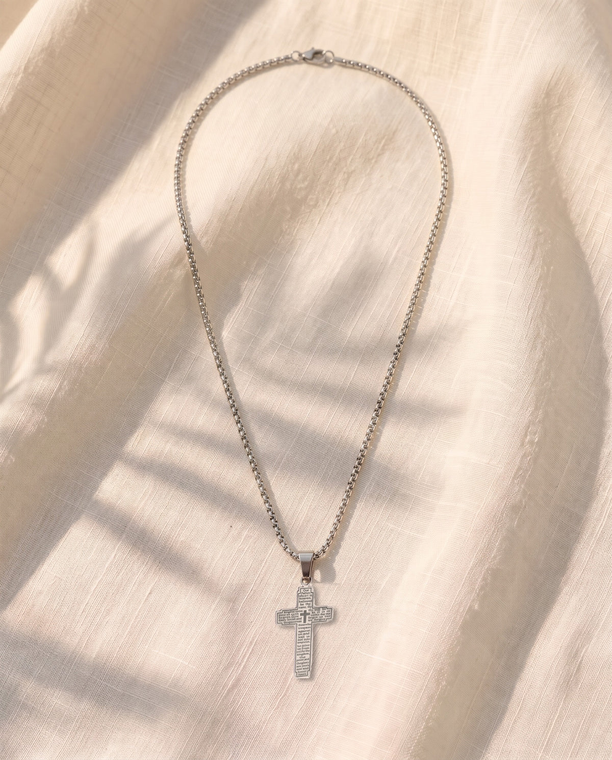Marco Maveri Lord's Prayer Cross Pendant Necklace laid on cream linen, Silver