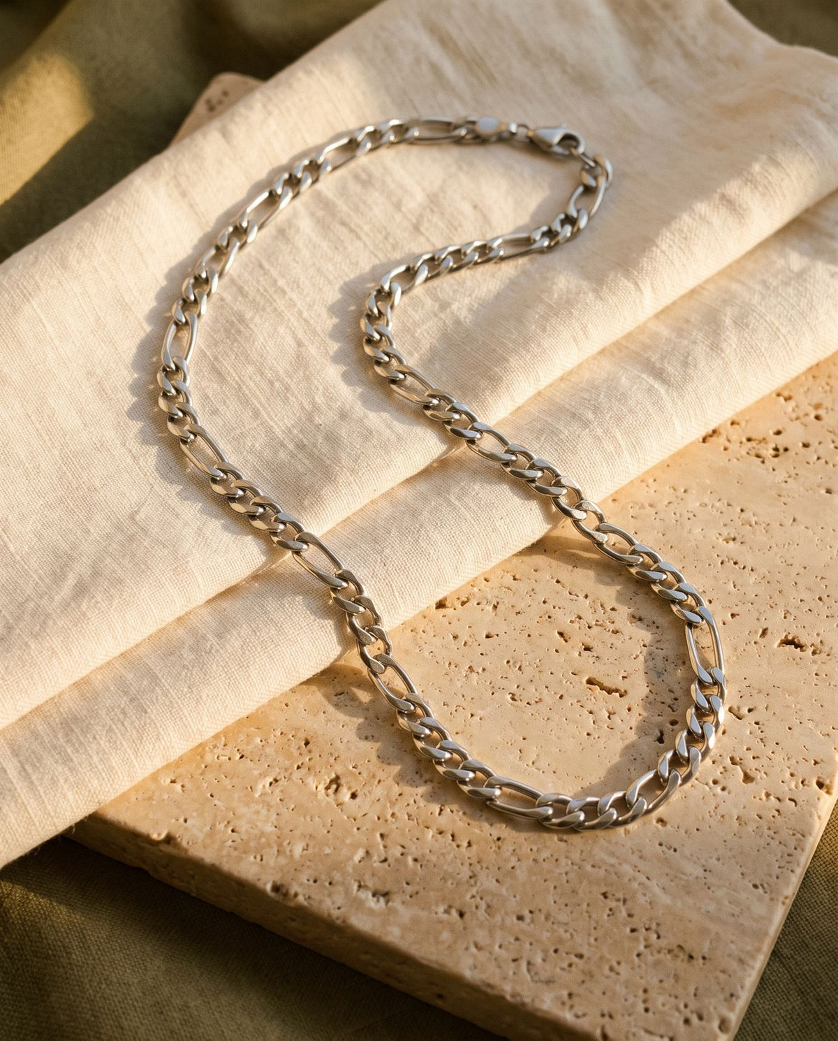 Figaro chain laid on ecru linen and travertine, lobster clasp visible