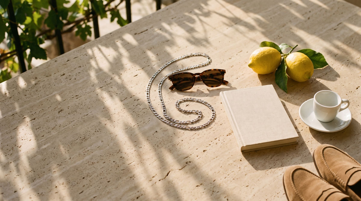 Figaro chain, sunglasses, lemons and a book on a travertine table under a pergola